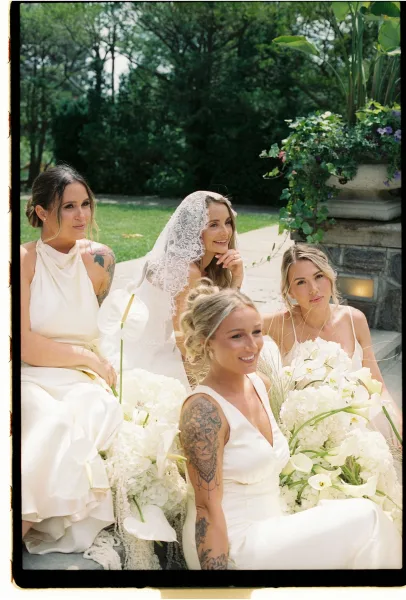 Bridesmaids portrait of an all-white bridal party sitting on stone steps, holding hydrangea and calla lily bouquets in a garden setting