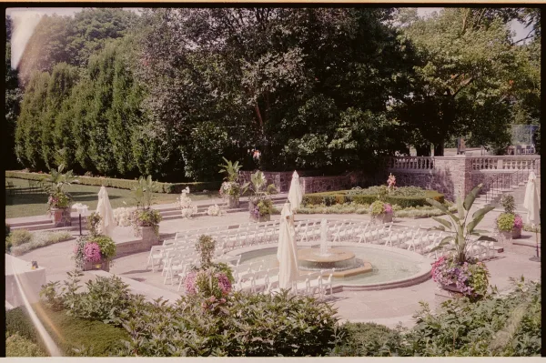 Outdoor ceremony setup with white folding chairs in a semicircle around a fountain, floral planters and umbrellas on a sunny garden terrace