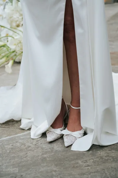 Bridal shoes, lace wedding heels with bow details and an ankle bracelet peeking beneath a white dress hem on a stone floor backdrop