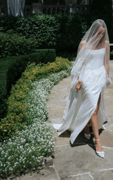Bridal portrait of a bride walking outdoors in a strapless satin gown with thigh slit, lace-edged veil over face on a sunlit stone walkway