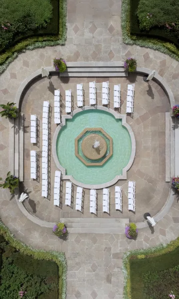 Ceremony setup with outdoor wedding ceremony seating of white folding chairs facing a fountain in a stone courtyard with steps and hedges
