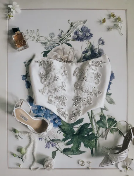 Bridal accessories flat lay with pearl necklace, beaded corset top, lace heels, rings and perfume on a floral print surface with orchid blooms