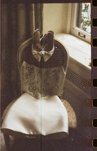 Wedding dress detail with beaded wedding dress bodice draped over an upholstered chair, bridal heels with ribbon ties by a window