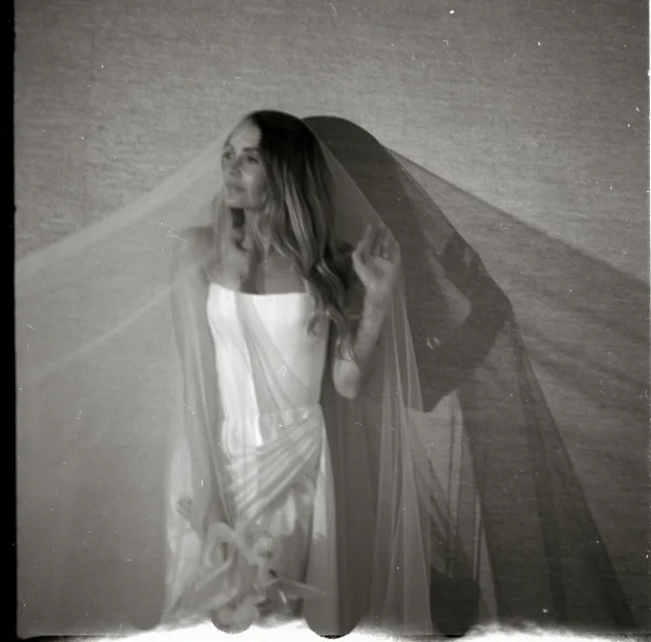 Bridal portrait in black and white with veil over her face, strapless dress and bouquet, posed by a plain wall with shadow