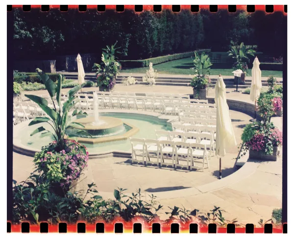 Ceremony setup with outdoor ceremony seating in a circular layout around a fountain, framed by white folding chairs and patio umbrellas on a stone terrace