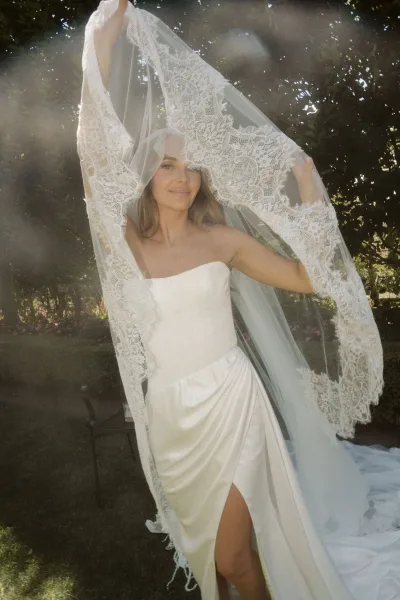 Bridal portrait of a bride in a strapless satin gown holding her lace veil overhead in a sunlit garden with trees and lawn