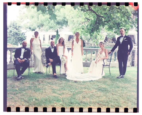 Wedding party portrait with bride, bridesmaids and groomsmen in tuxedos, bridal veil and bouquet on a lawn beneath trees by stone balustrade