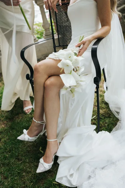 Bridal portrait of a bride sitting in chair, legs crossed, showing lace garter as an orchid bouquet rests on her lap in a garden lawn setting