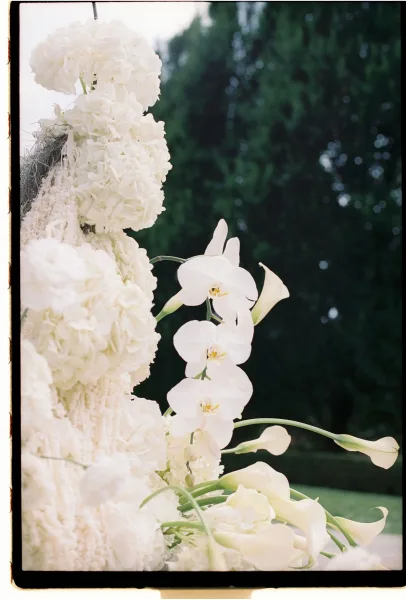 Wedding floral arrangement of white wedding flowers with hydrangeas, orchids, and calla lilies hanging above a lawn altar framed by evergreens