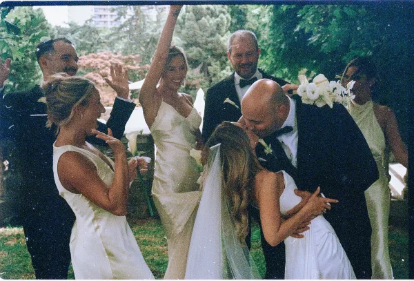 Wedding kiss moment as groom dips bride in veil and gown, bridesmaids cheer with bouquet and champagne in outdoor garden setting