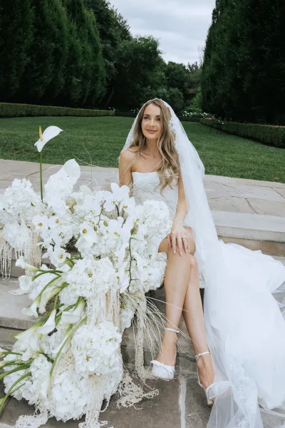 Bridal portrait of bride sitting on stone steps in a strapless wedding dress with lace veil, holding all-white calla lily bouquet in garden greenery