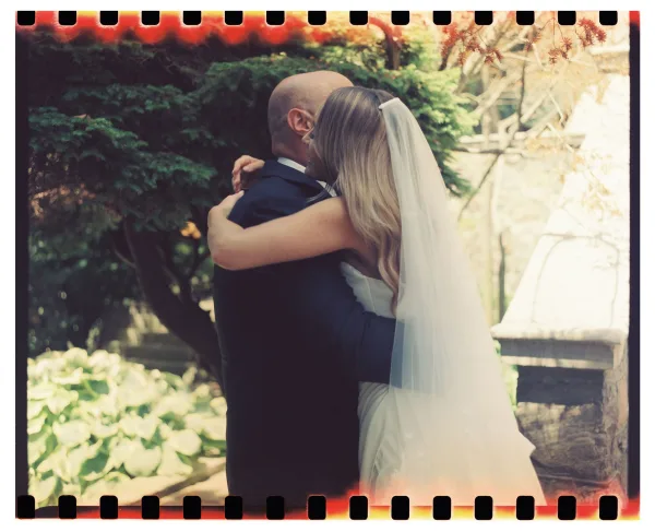 Wedding first look as the bride hugging groom in a sunlit garden, her long bridal veil and strapless dress against his black suit boutonniere