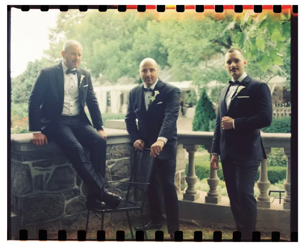 Groomsmen portrait of three men in black tuxedos with bow ties and boutonnieres, standing and sitting on a stone terrace railing by trees