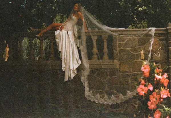 Bridal portrait with long wedding veil draped over a stone balustrade, bride in strapless satin gown with lace-trim veil edge amid garden trees and pink flowers