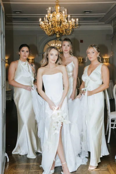 Bridal party portrait of bride with bridesmaids in champagne satin dresses, holding veils and bouquet beneath a chandelier in paneled hallway
