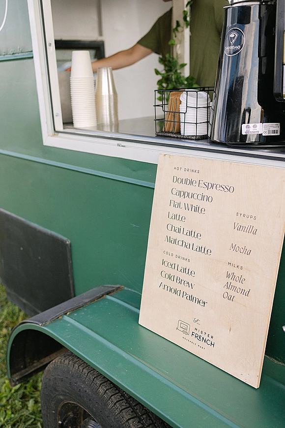 Coffee cart menu at a wedding coffee cart service window with espresso machine, paper cups, and stir sticks on a green trailer on grass lawn