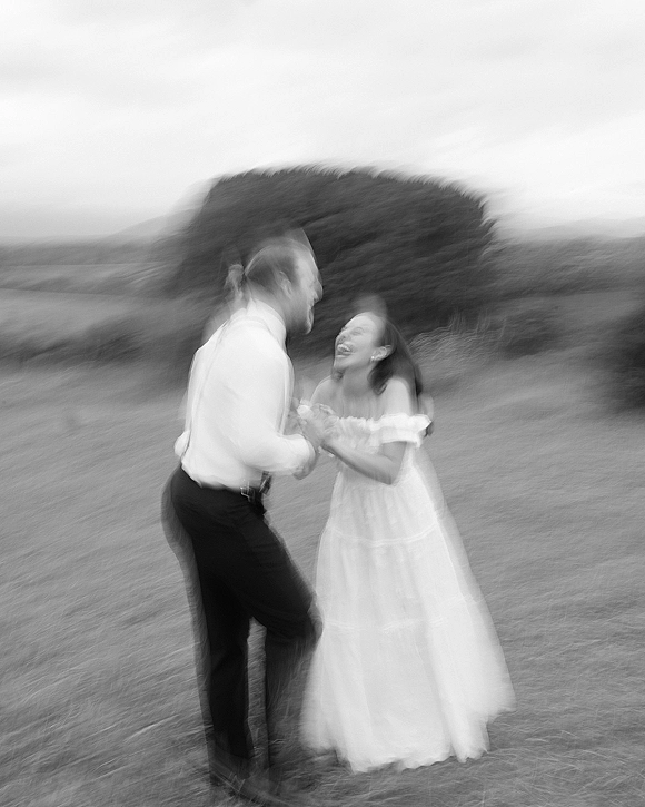 Wedding couple portrait in a black and white wedding photo, bride in off-shoulder gown and groom in suspenders laughing in a grassy field
