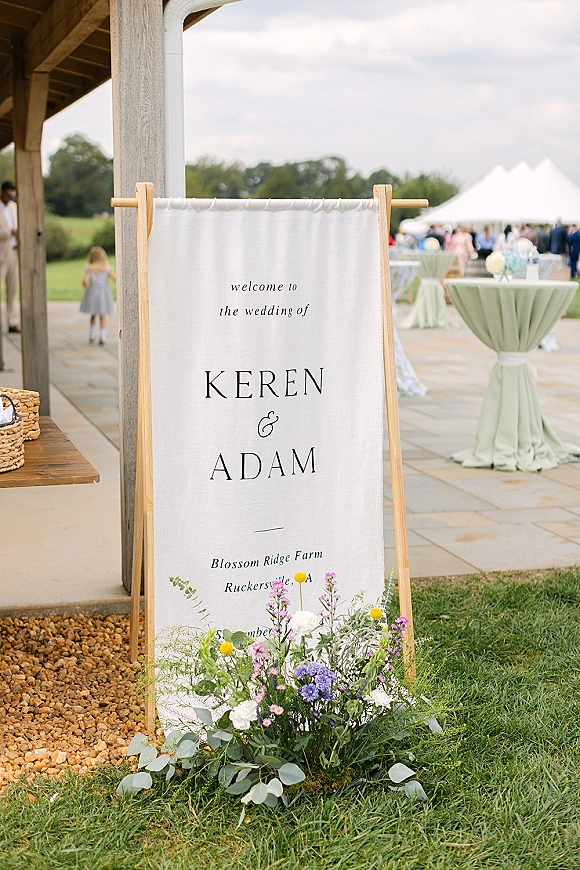Wedding welcome sign on a wooden stand with a linen wedding sign banner, greenery and florals at an outdoor patio tent reception entrance