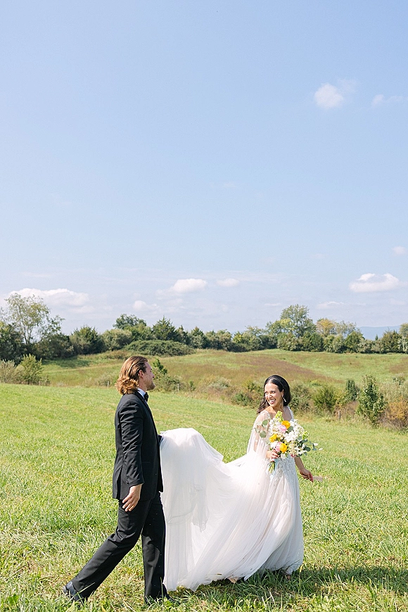 Couple portrait in an outdoor wedding portrait, bride and groom walking and laughing as her long veil trails across a grassy hill under blue sky