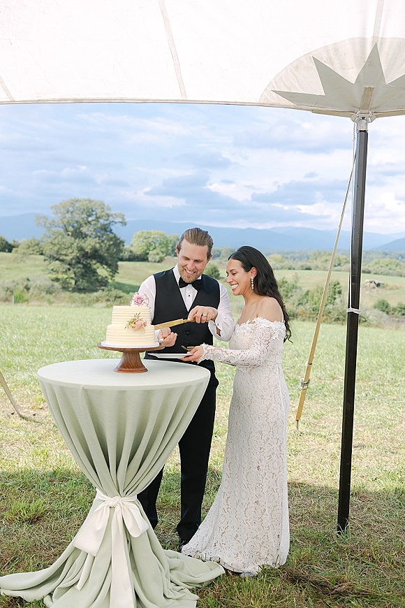 Cake cutting by bride and groom cutting cake at a sweetheart table under a tent canopy, with mountains behind and a floral-topped cake stand