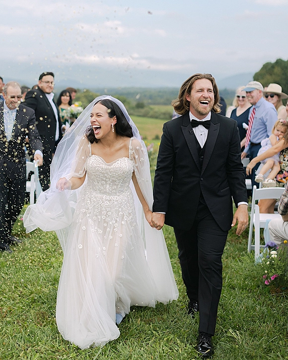 Wedding recessional as bride and groom walk hand in hand through confetti, guests cheering beside white chairs on a mountain lawn