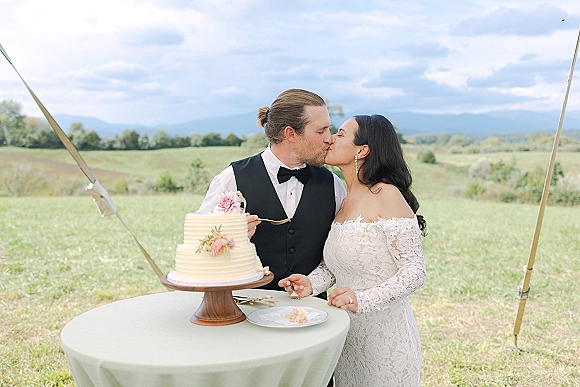 Wedding cake cutting as the bride and groom slice a buttercream cake with floral greenery accents under a tent in rolling hills