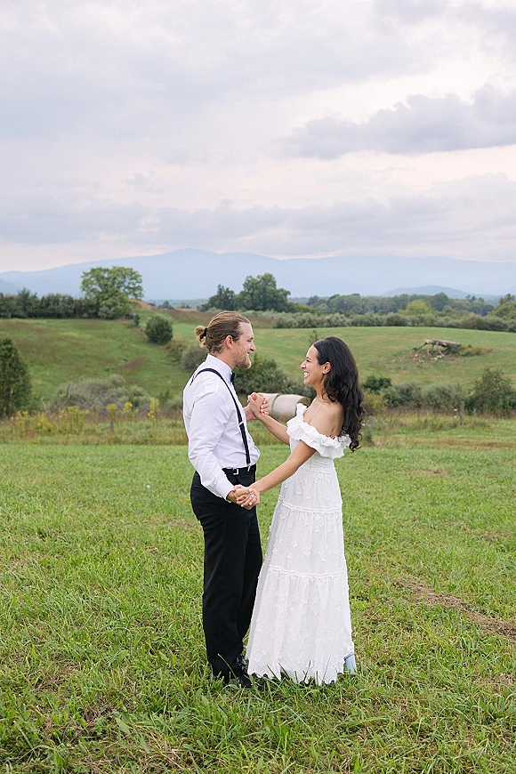 Couple portrait of bride and groom holding hands, her off-shoulder gown and his suspenders and bow tie in a meadow with mountains under clouds