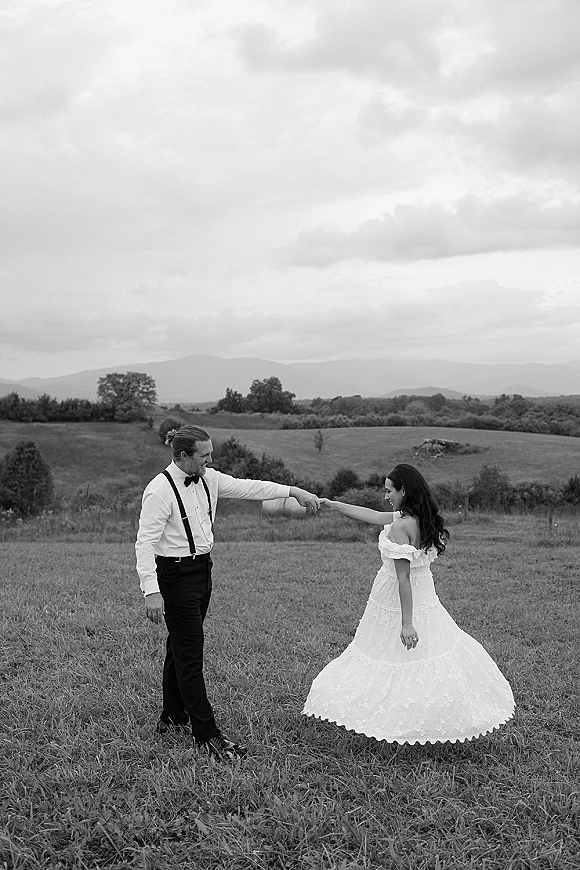 Couple portrait in a black and white wedding photo, bride in full skirt dress twirling as groom in suspenders holds her hand in a mountain field