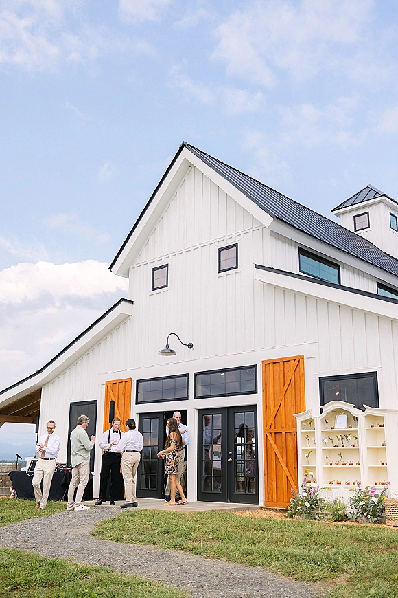 Wedding venue exterior of a white barn wedding venue with wood barn doors and black glass doors, gravel path, lawn, and blue sky