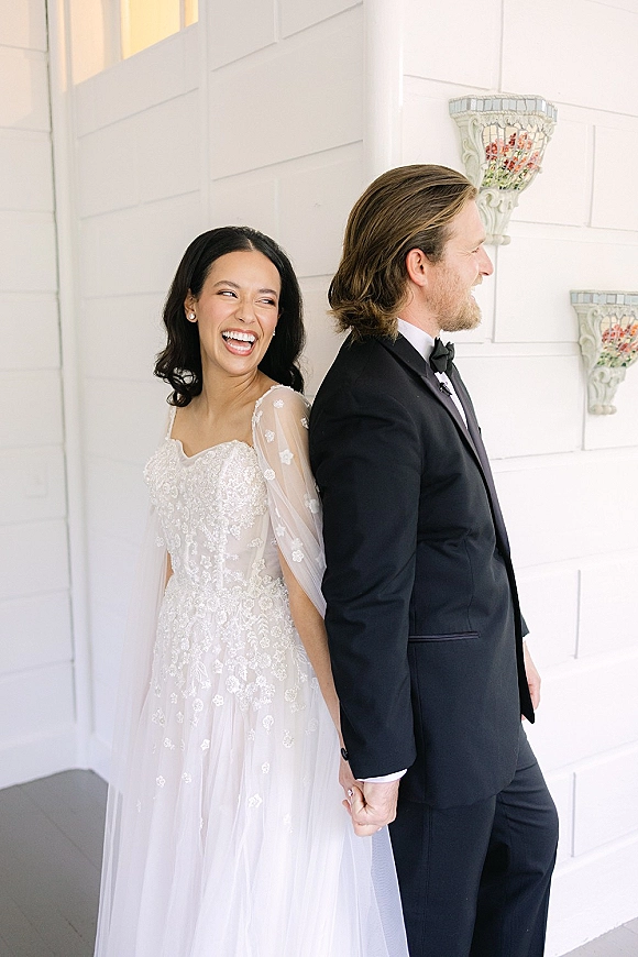 Wedding couple portrait with bride and groom holding hands, laughing back to back by a white paneled porch wall, lace sleeves and black tuxedo
