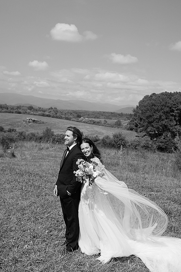 Couple portrait in black and white wedding portrait style, bride hugging groom as her long veil blows, bouquet in a meadow with hills and mountains