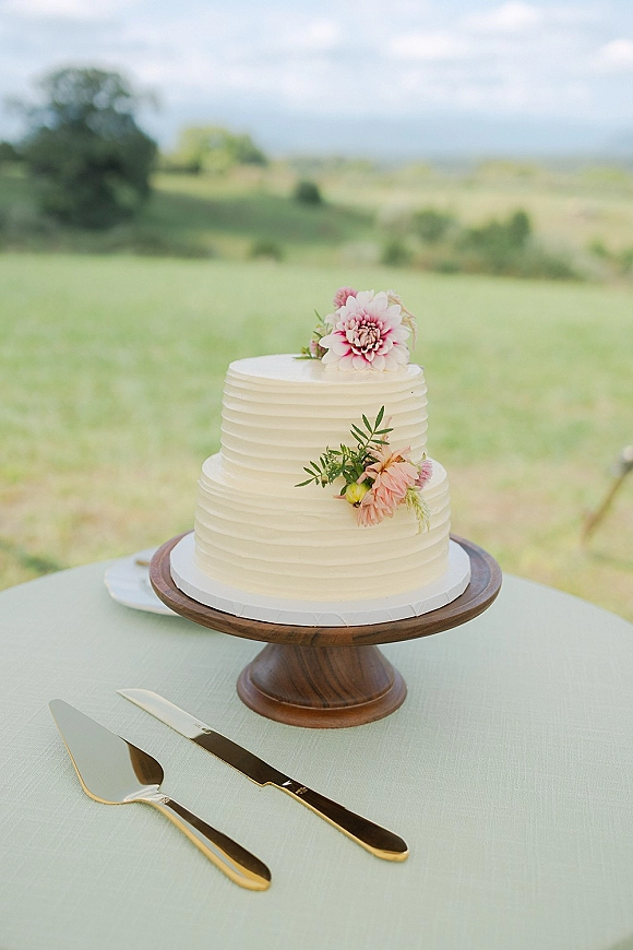 Wedding cake with two tier buttercream finish topped with fresh flowers and greenery on a stand, set outdoors with rolling hills backdrop