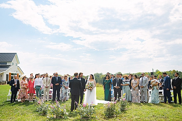 Wedding ceremony outdoors with bride in veil and groom in black suit on a white aisle runner, guests standing near a barn on grass lawn