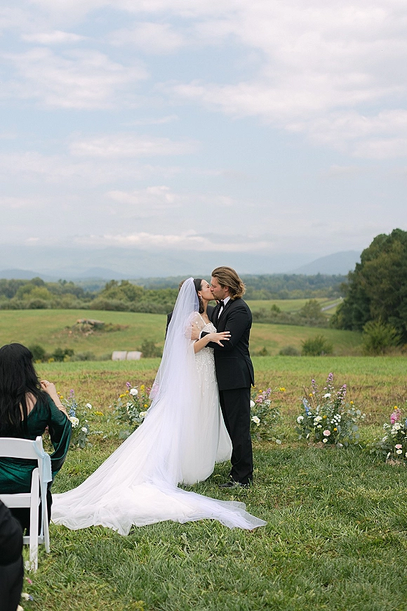 Wedding kiss as bride in lace gown with veil and long train meets groom in tuxedo, framed by meadow chairs and distant mountains