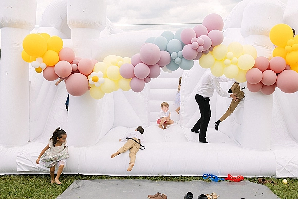 Wedding bounce house with pastel balloon garland and daisy balloons on a grass lawn under a cloudy sky, set as a playful backdrop