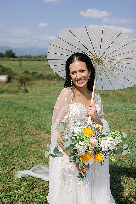Bridal portrait of a bride holding a parasol and bouquet in a lace wedding dress, standing in a meadow with hay bale and distant hills