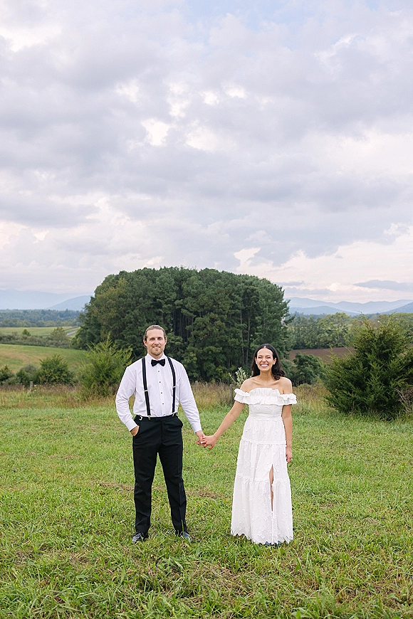 Couple portrait of bride and groom holding hands in a meadow with rolling hills and distant mountains under a cloudy sky, bow tie and suspenders