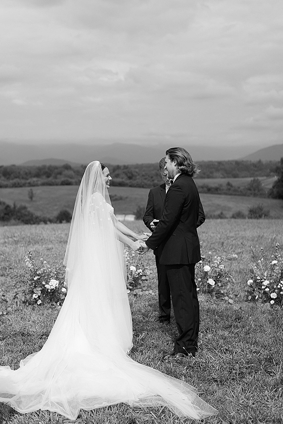 Wedding vows during an outdoor wedding ceremony as bride and groom hold hands, veil trailing in a wildflower meadow with mountains under clouds