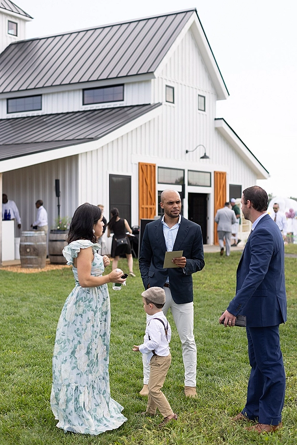 Wedding guests mingling at cocktail hour outside a white barn, chatting on the lawn with drink bottle accents near wooden barrels