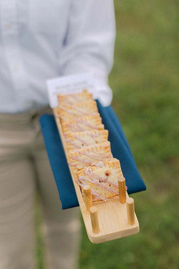 Wedding dessert tray of bite-size pastry slices on a wooden serving board with a place card and linen napkin outdoors on grass lawn