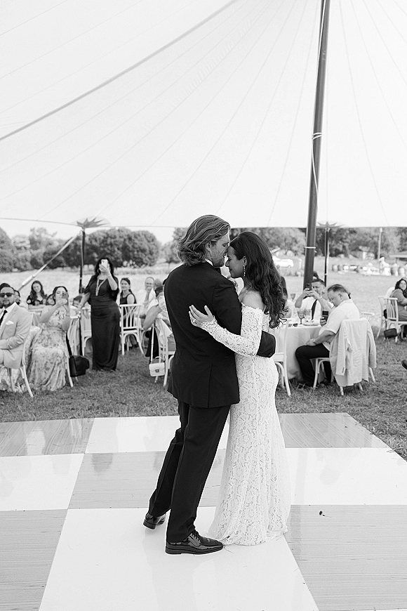 First dance wedding first dance photo of bride in lace gown and groom in suit slow dancing on floor under tent as guests watch on lawn