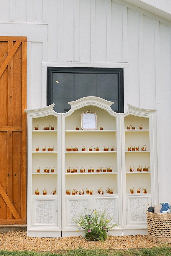 Wedding favor display with honey wedding favors in mini glass jars on white cabinet shelves, framed sign and wildflower accent by barn door