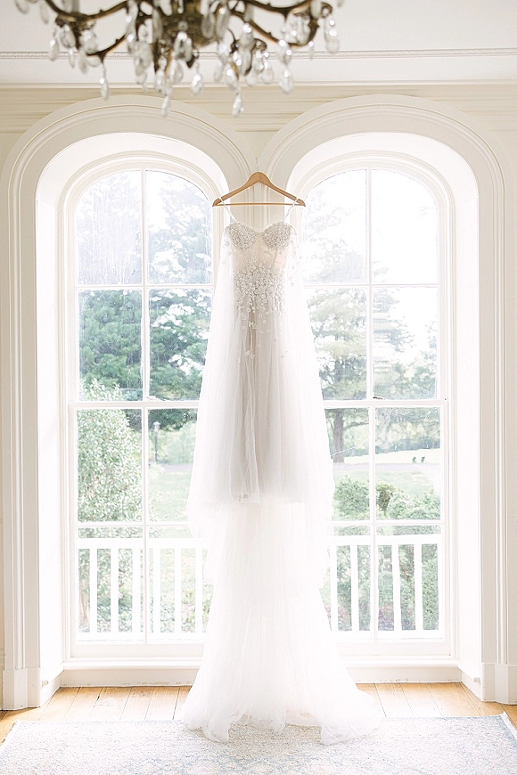 Wedding dress with lace bodice hanging on a hanger in front of arched windows, tulle skirt softly lit with a chandelier overhead