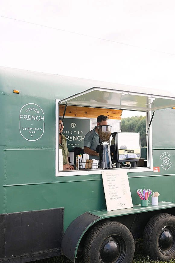 Wedding coffee cart espresso bar wedding setup with espresso machine, grinder, menu sign, and paper cups under an awning in a field