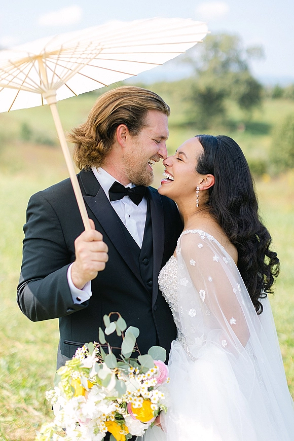 Couple portrait of bride and groom laughing under a white parasol, her bouquet in hand, in a meadow with rolling hills behind