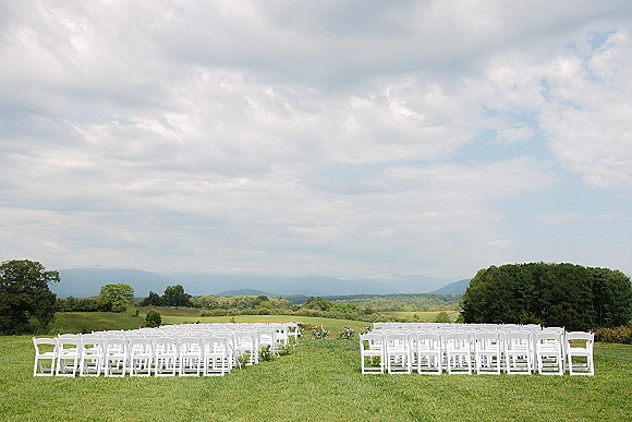 Ceremony seating with white folding chairs and low aisle floral arrangements on a grass lawn, with mountains and trees under a cloudy sky