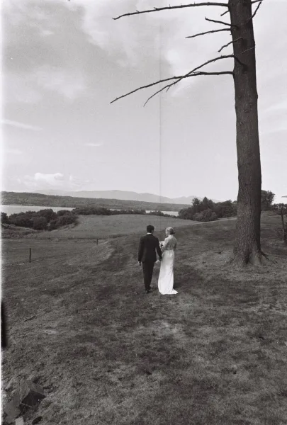 Couple portrait of bride and groom holding hands, walking away in a meadow toward a lake and mountains under a cloudy sky