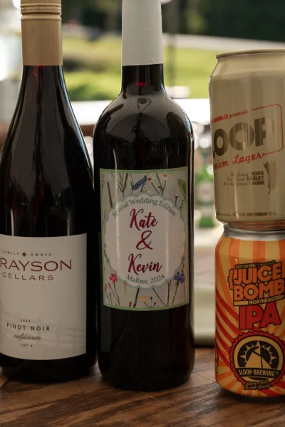Custom wedding wine label on bottles with personalized wine bottle label details beside beer cans on a wooden table, blurred lawn greenery behind