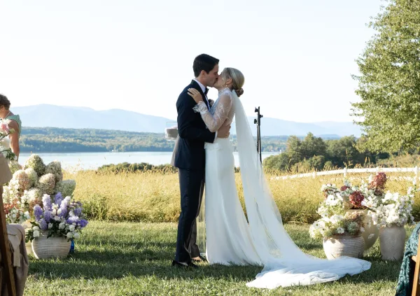 Wedding kiss at an outdoor wedding ceremony, bride in lace sleeves and long veil with groom in tuxedo by lakeside mountains and florals