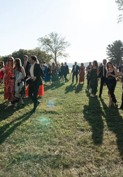Wedding guests in formal dresses and suits wearing sunglasses walk between chair rows on a grassy lakeside lawn with trees and hills